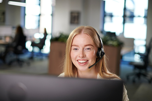A smiling support agent wearing a headset at her desk in an open-plan office