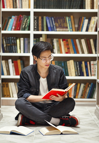 A student sitting cross-legged in front of a bookshelf, reading a book — explore the docs