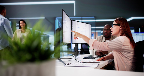 A woman pointing at her monitors while colleagues look on in a modern office